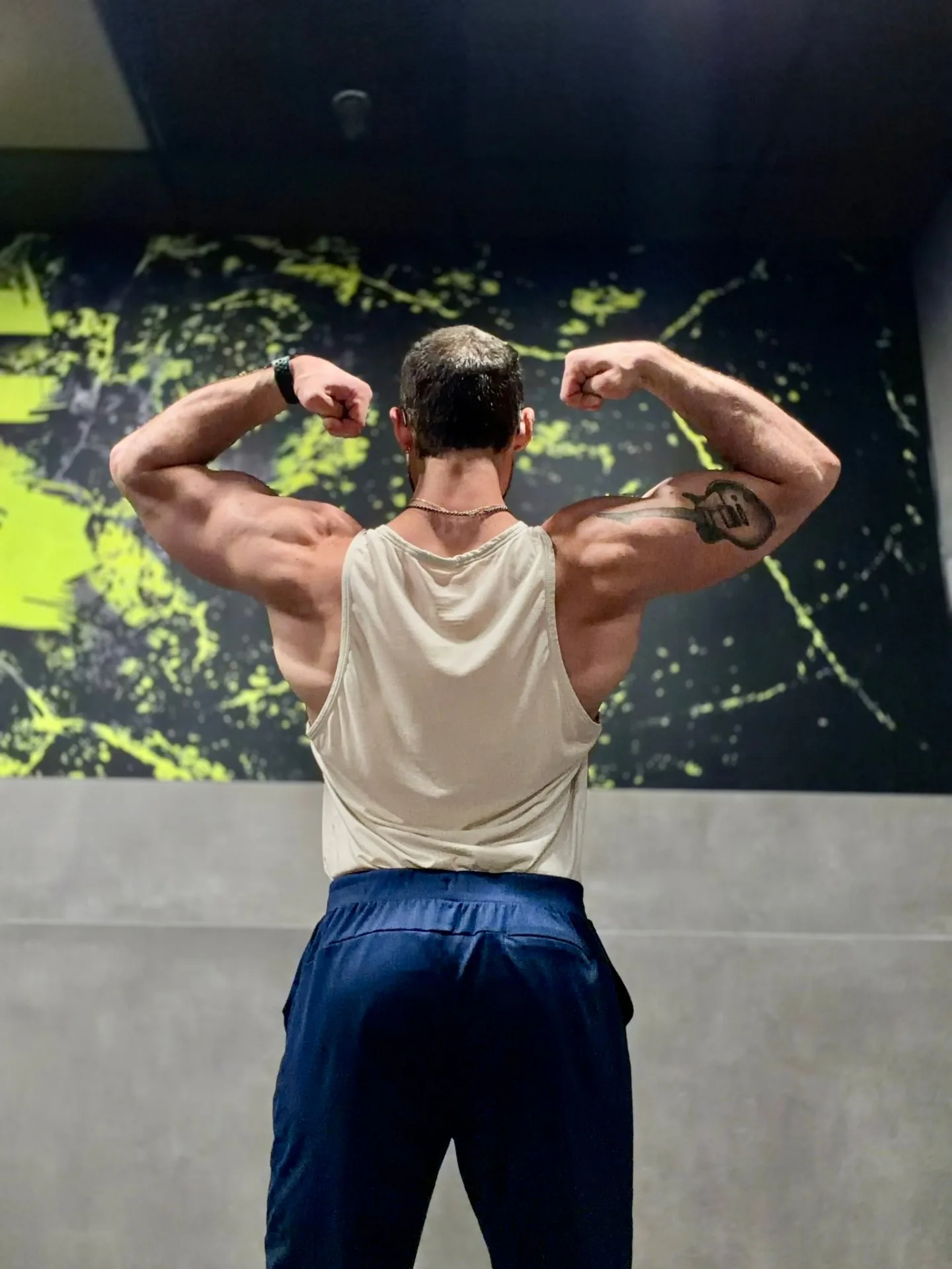 The Anabolic Architect coach holding a rear double-biceps pose, lit from above against a dark studio backdrop