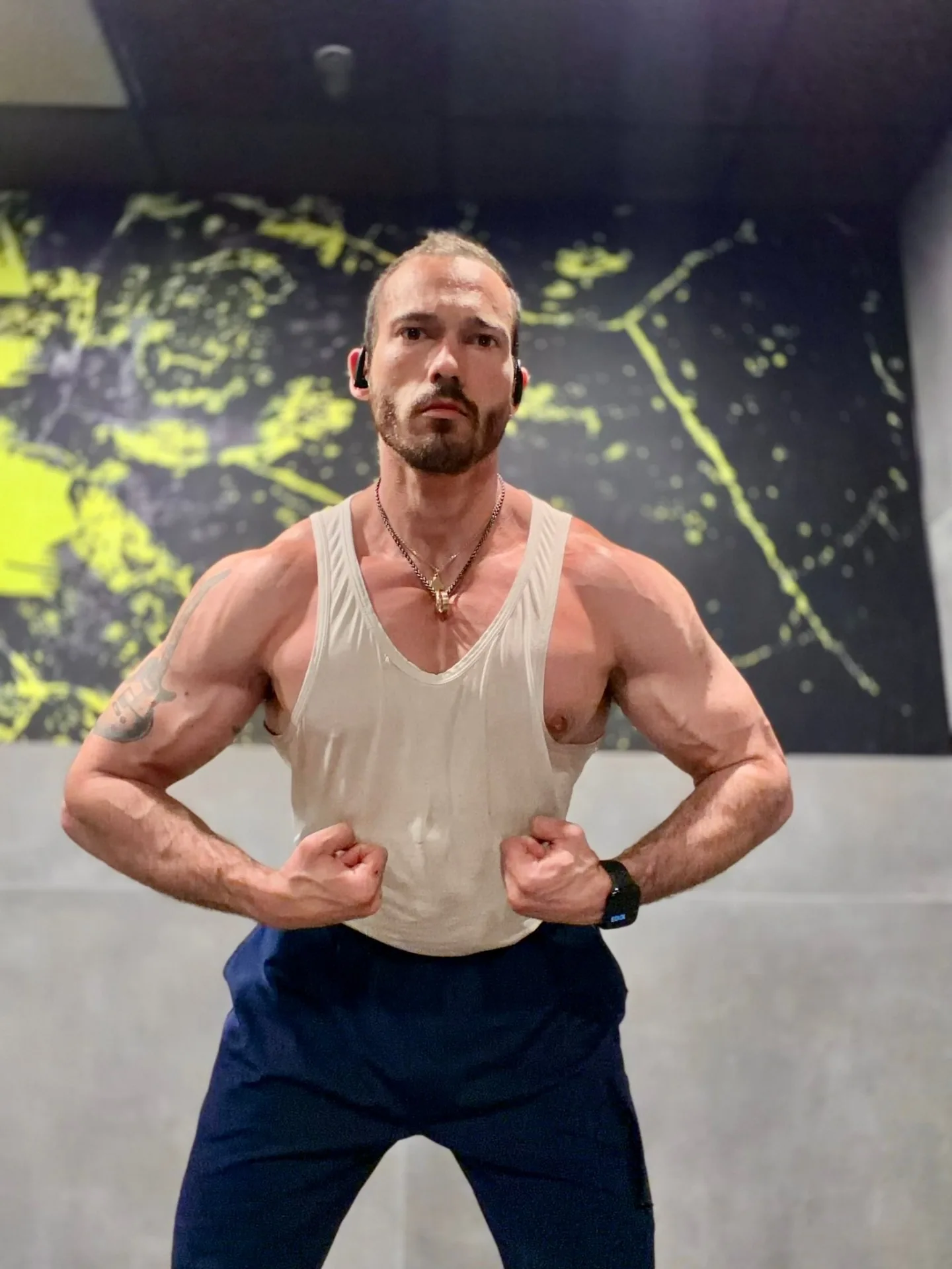 The Anabolic Architect, founder and head coach, holding a front double-biceps pose in a dim industrial gym